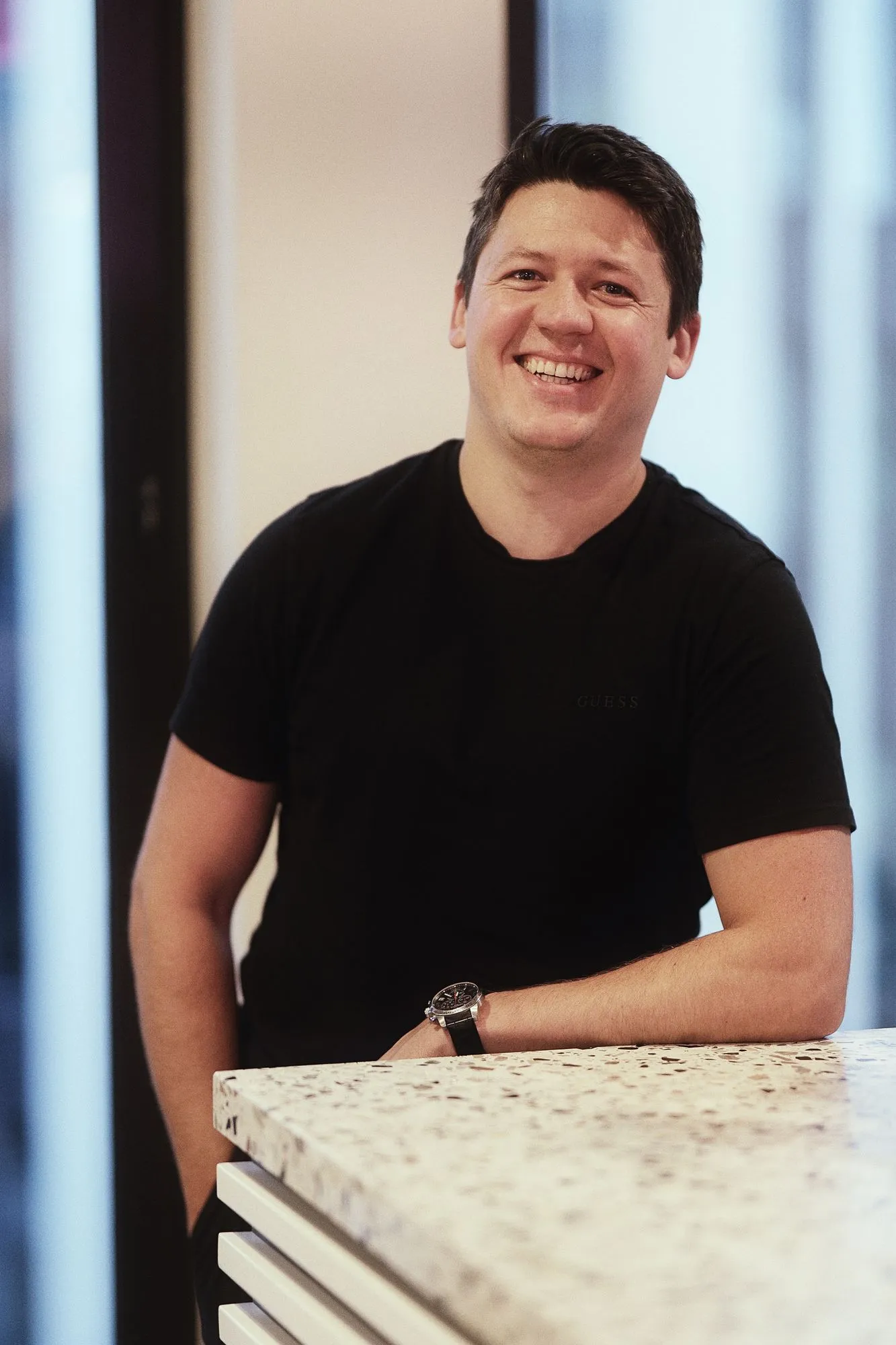 Alex Kukharenko leaning on a counter and laughing, in a black t-shirt.